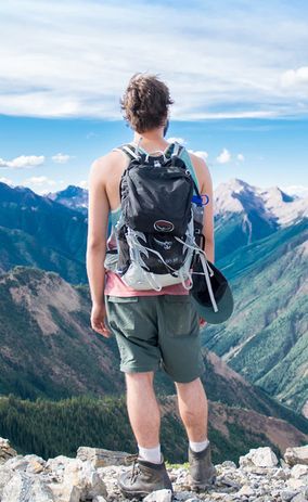 Турист со рюкзаком стоит спиной на вершине горы и смотрит на горный хребет под голубым небом.