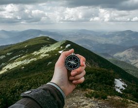 Рука с компасом на фоне горного хребта под пасмурным небом.