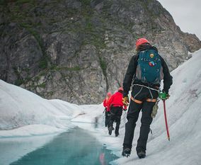 Группа альпинистов в яркой одежде и касках идет по леднику рядом с бирюзовым ледниковым озером.