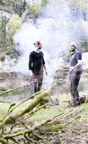 Туристы на привале разводят костер на берегу реки в лесу.